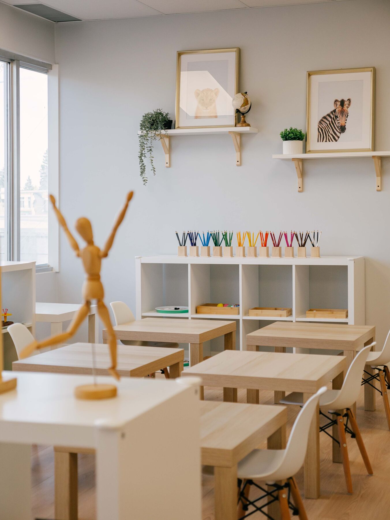 A tidy classroom with cleared table tops, pots of coloured pencils lined up on a shelf, and framed prints on the wall.