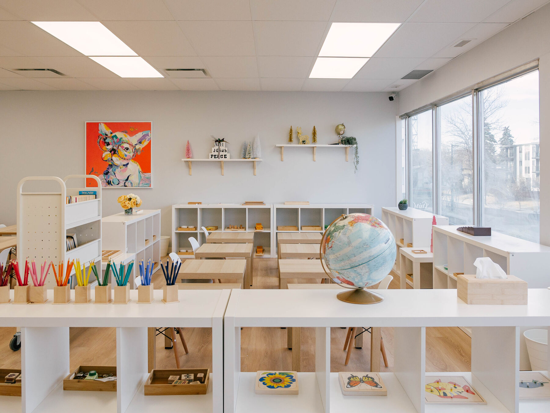Open classroom shelving holding activity trays, a globe and pots of coloured pencils sorted by hue.