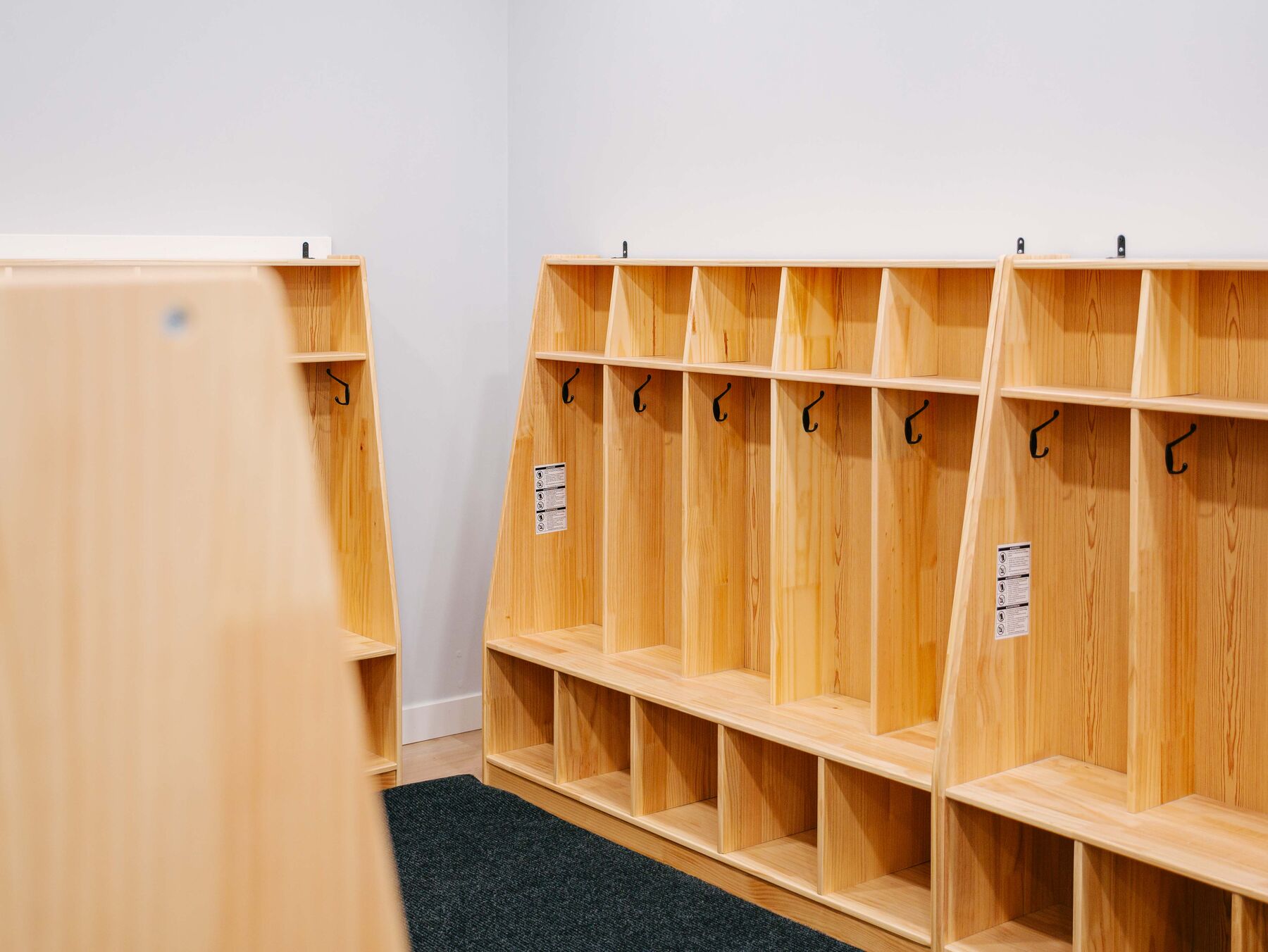 Wooden cubbies along a wall above dark hard-wearing carpet, with a bench edge in the foreground.