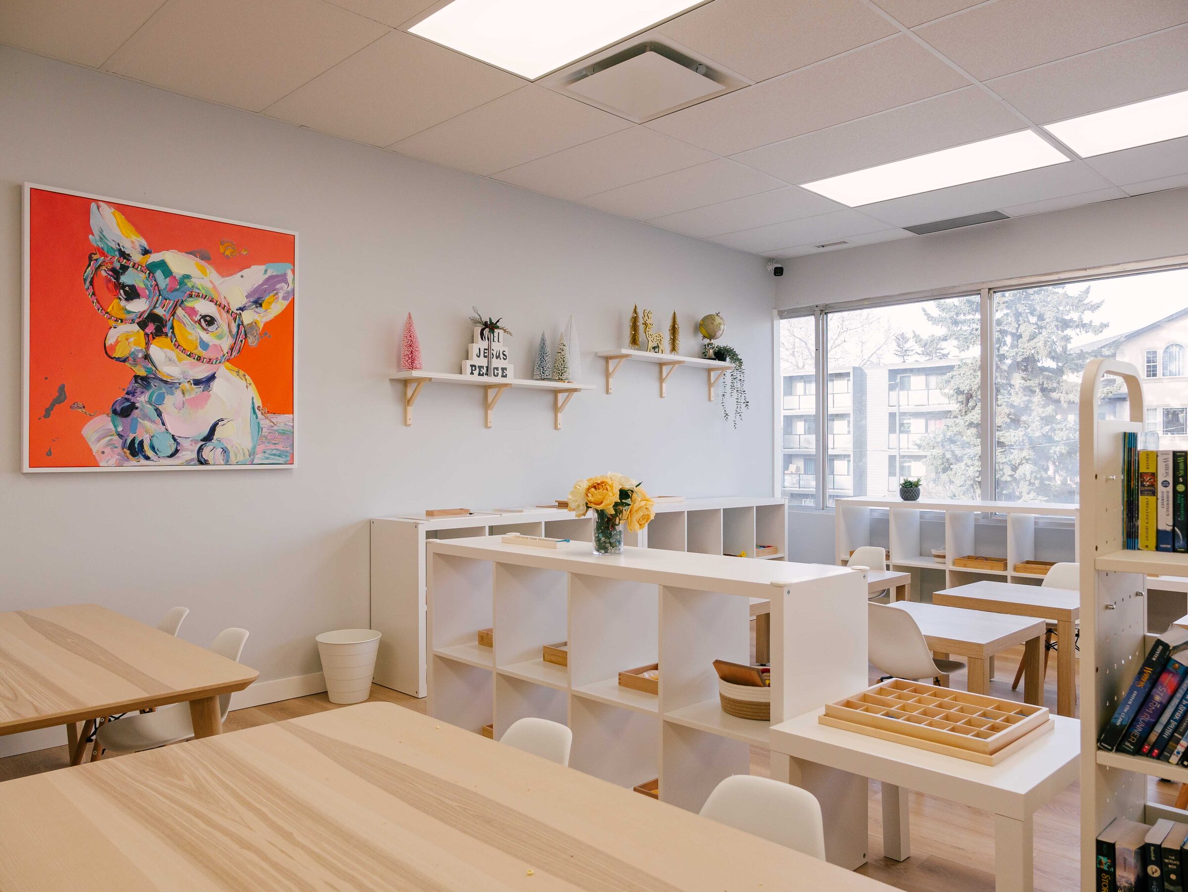 A Mindful Playful classroom seen from the doorway: low white shelving, pale work tables, and a wall of windows.