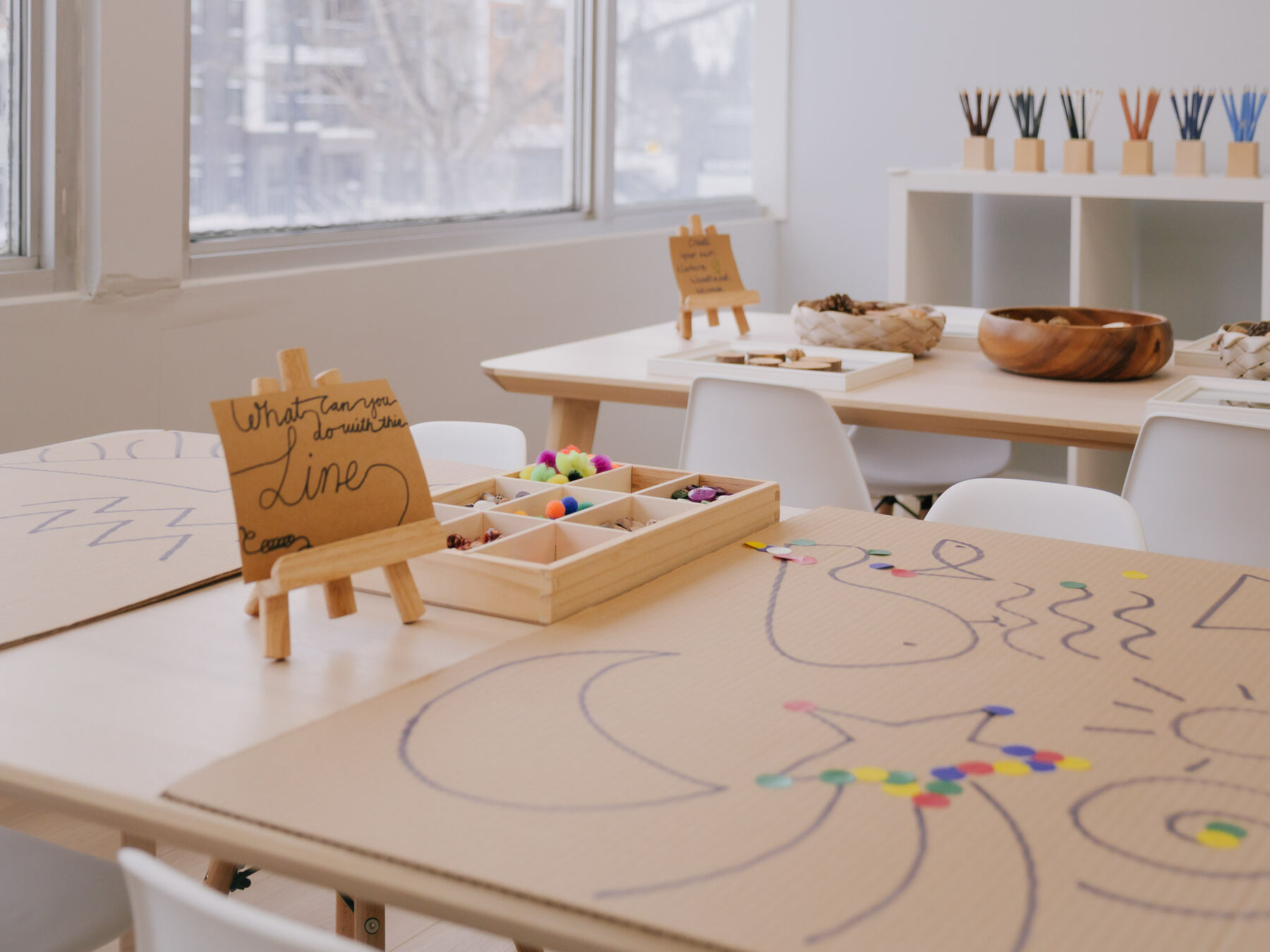 Two studio tables covered end to end with paper the children have drawn on, with child-sized chairs and a bowl of natural materials behind.