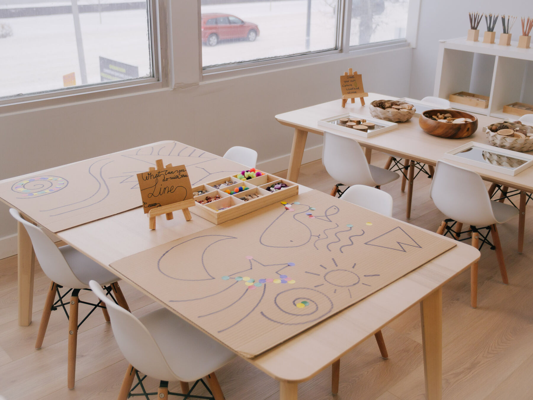 A studio table covered in paper with a hand-lettered card on a small easel reading 'What can you do with the line?', beside a compartment tray of pom-poms and beads.