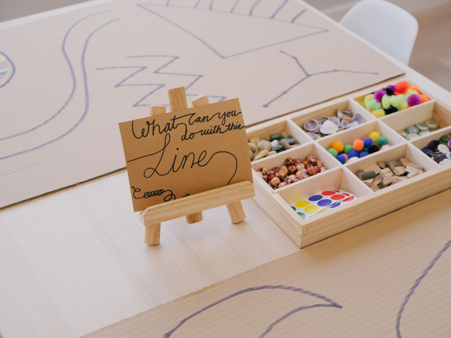 A wooden compartment tray of pom-poms, beads, buttons and stones resting on paper marked with drawn lines.