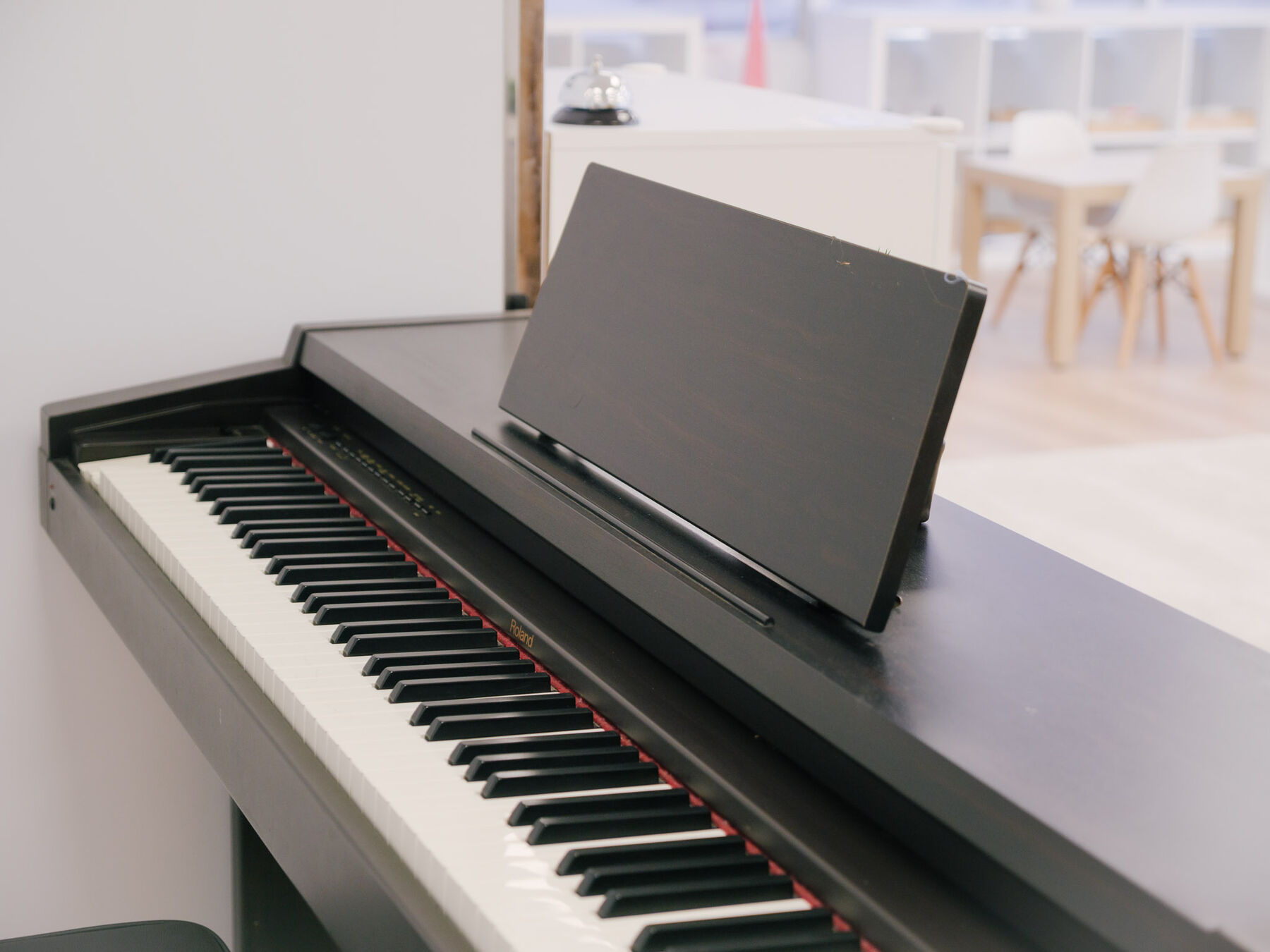 An upright digital piano with its music stand raised and keys uncovered, standing at the edge of a classroom.