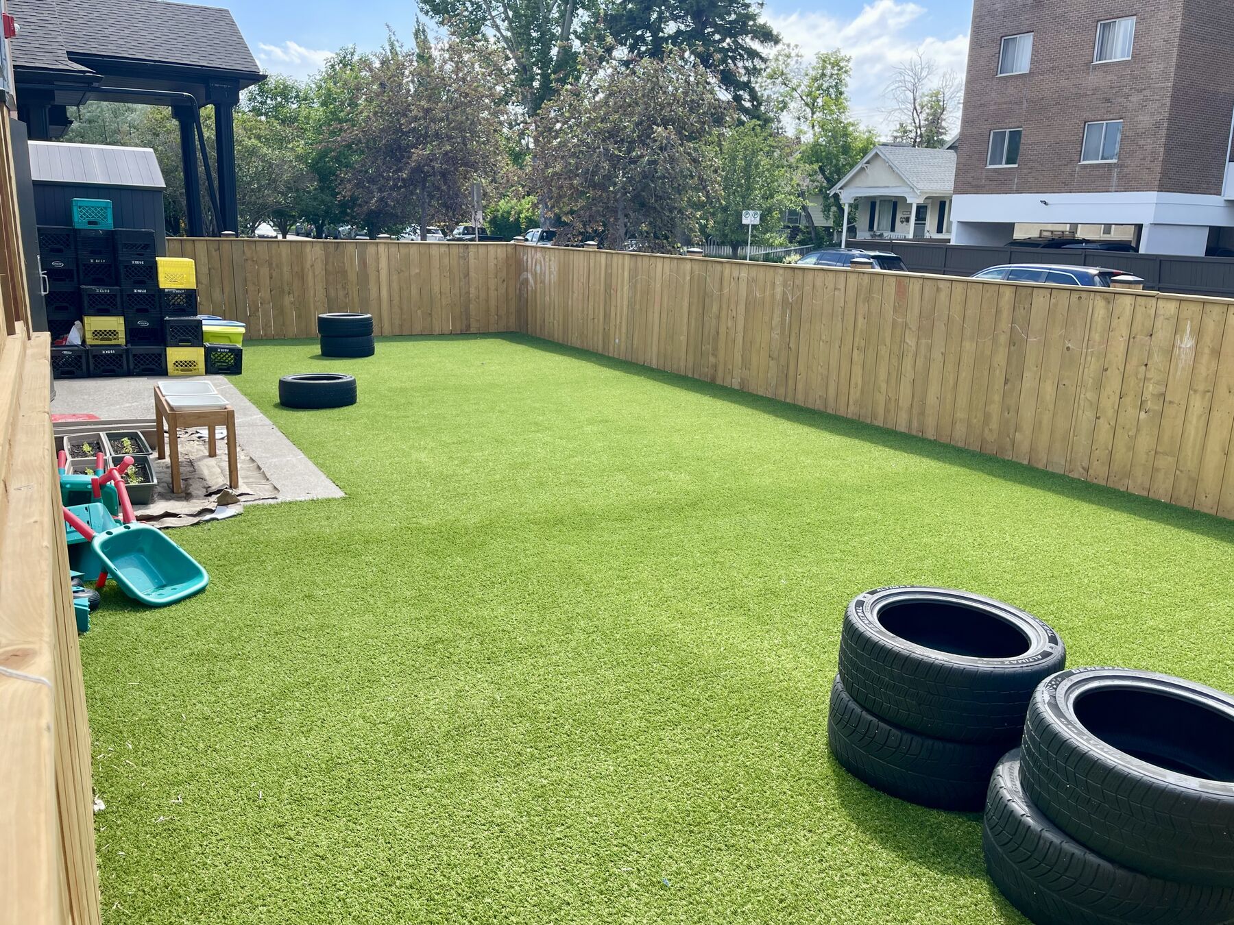 A long stretch of green turf in a fully fenced yard, with a wheelbarrow and play equipment at the far end.
